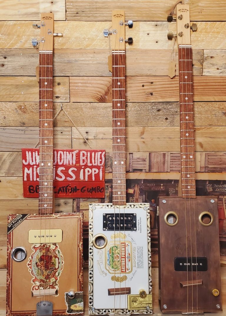 Guitare cigar box cigarboxmusic.fr Trois guitares cigar box artisanales en bois, décorées avec des motifs distinctifs et des étiquettes.